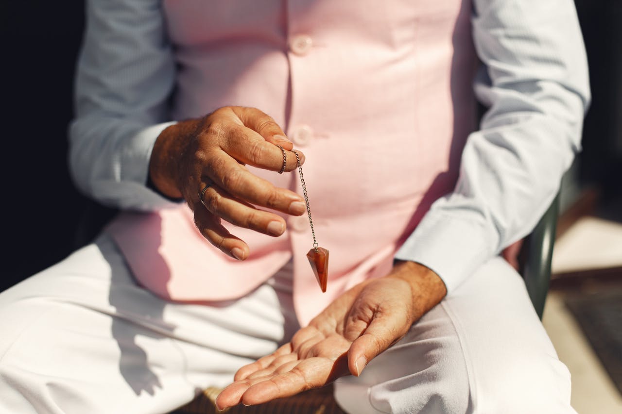 A man in soft pastel attire holding a gemstone pendulum over his palm outdoors.